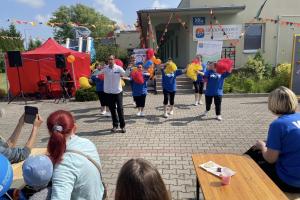 Na zdjęciu widać grupę tańczących osób z kolorowymi pomponami. W tle zielony budynek Środowiskowego Domu oraz czerwony namiot, pod którym jest didżej. Na pierwszym planie widać kawałek stołu i parę osób oglądających występ.