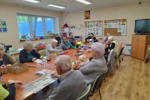 Pomieszczenie , na środku stoi wielki stół przy którym siedzą seniorzy. Panie i panowie zajęci są  układaniem różnych układanek