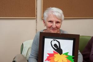 na zdjęciu widnieje uśmiechnięta pani. Prezentuje swoją pracę, wyklejankę- baletnicę, która ma spódnicę zrobioną z kolorowych liści