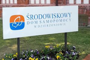 tablica informacyjna przed budynkiem Ośrodka , pod nią znajduje się rabata pełna kwiatów