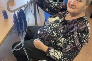 na końcu stołu siedzi na krześle kobieta, w ręce trzyma pracę zrobioną z makramy- chmurkę i liście uplecione z niebieskiego sznurka
