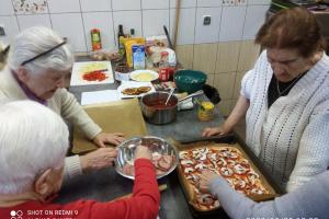 grupa Pań nakłada na pizzę składniki. Na blacie koło kobiet widać produkty użyte to przygotowania pizzy