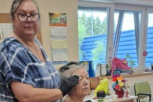 Na pierwszym planie terapeutka wacikiem zmywa twarz seniorki. seniorka siedzi przy stole