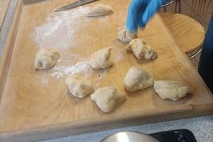 zdjęcie przedstawia leżącą na stole stolnicę. Oprószona jest ona mąką, leża na niej kawałki ciasta, z którego będą robione pączki. Obok stolnicy znajduje się waga elektroniczna,.