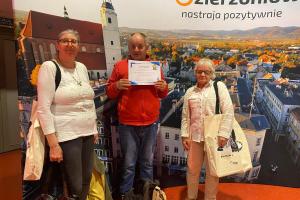 trzy osoby, dwie panie i pan pośrodku pozują do zdjęcia na tle plakatu panoramy Dzierżoniowa. W prawym górnym rogu plakatu umieszczony jest napis: Dzierżoniów nastraja pozytywnie