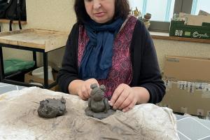 na zdjęciu widać panią, która siedzi przy stole. na stole leży materiał. Kobieta lepi z gliny figurkę