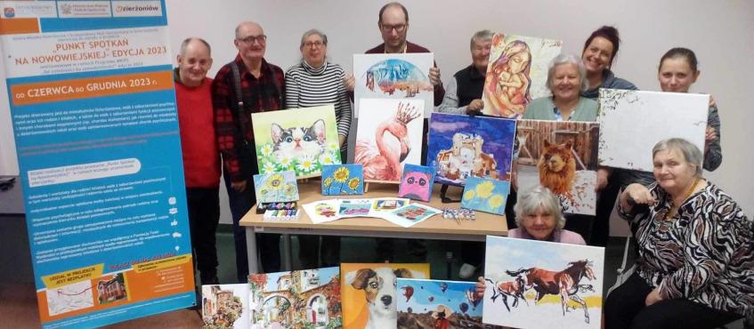 Grupa osób stoi i prezentuje obrazy, które namalowali podczas zajęć malarskich. Obok nich jest baner informujący o projekcie.