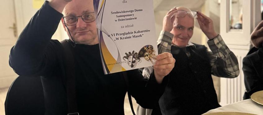 Dwóch panów siedzących za stołem pozuje do zdjęcia, jeden z nich trzyma dyplomm