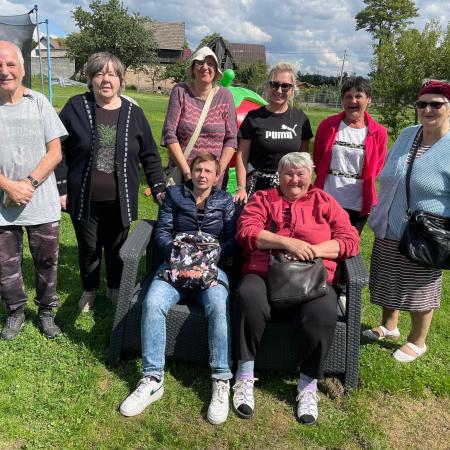 Wyświetl większy obrazek zdjęcie prezentuje grupę ludzi. Po obu stronach stoją trampoliny. Dwie kobiety siedzą na krzesłach. Wokół widać zabudowania, kilka drzew i pogodne niebo