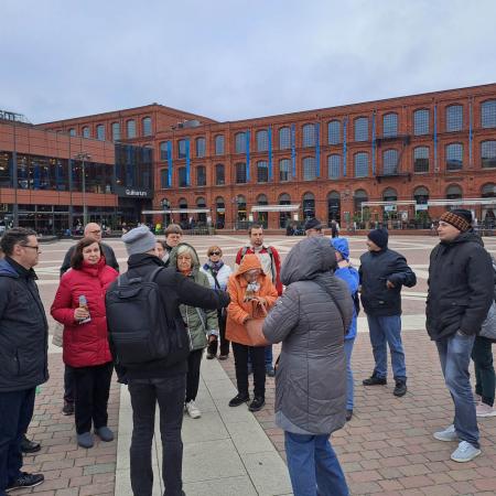 Wyświetl większy obrazek Wybrukowany kostką plac. W tle budynki manufaktury z czerwonej cegły. Na placu stoi grupa ludzi w kurtkach, słuchają przewodnika.