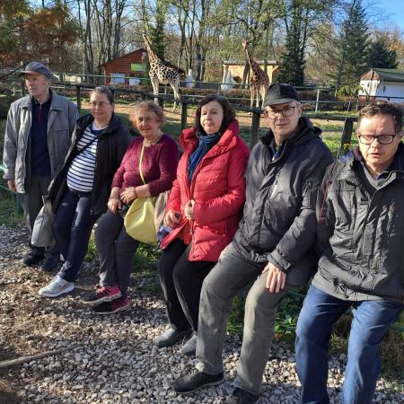 Wyświetl większy obrazek grupa ludzi, trzech mężczyzn i trzy kobiety siedzą na barierce przed wybiegiem dla żyraf. W tle chodzą dwie żyrafy