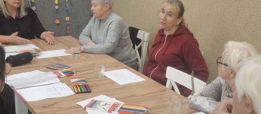 Uczestnicy grupy wsparcia siedza przy wielkim stole. Przed sobą mają kartki